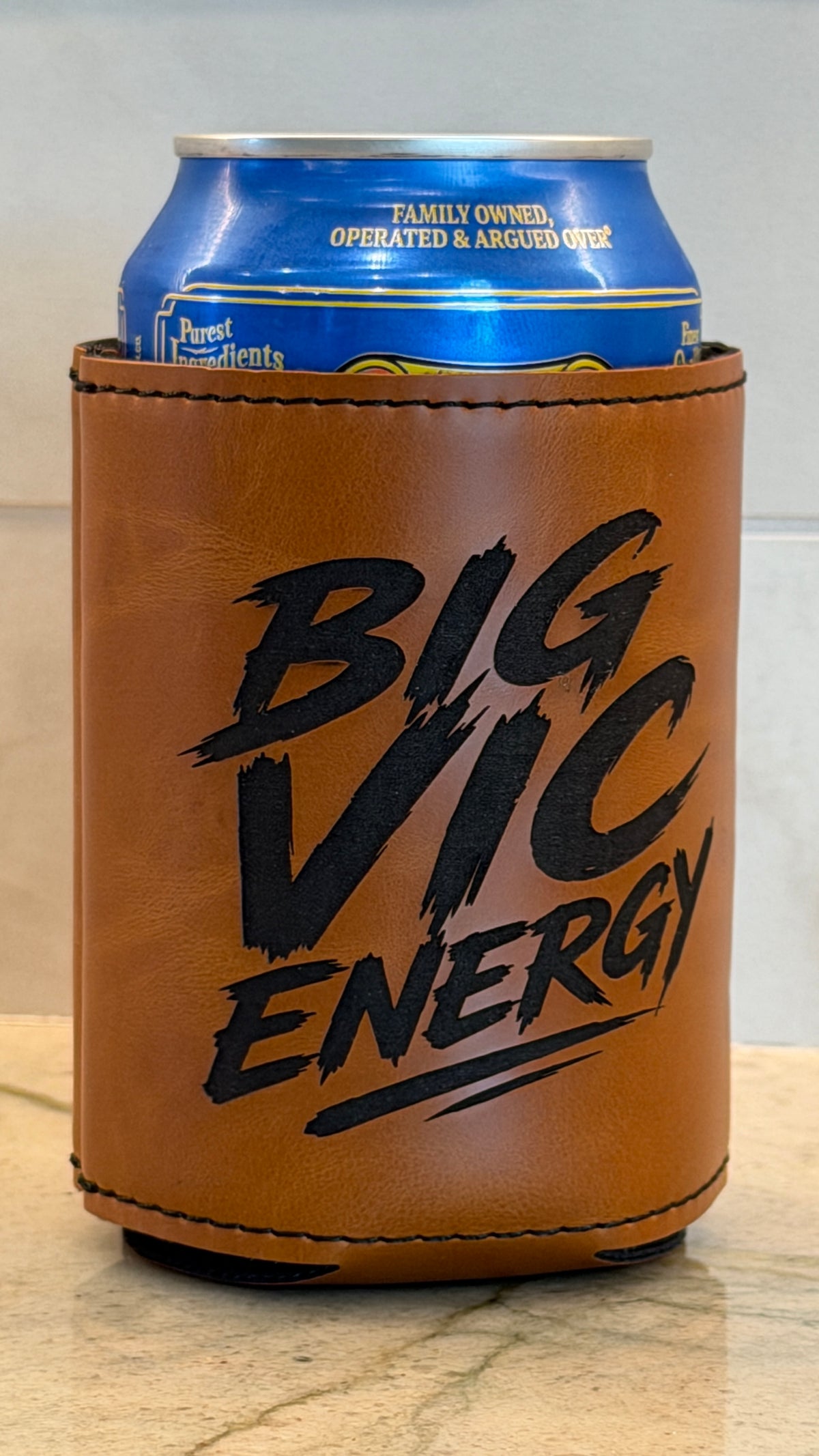 A brown leatherette can cooler sleeve with black 'BIG VIC ENERGY' text, holding a blue beverage can on a light stone surface.