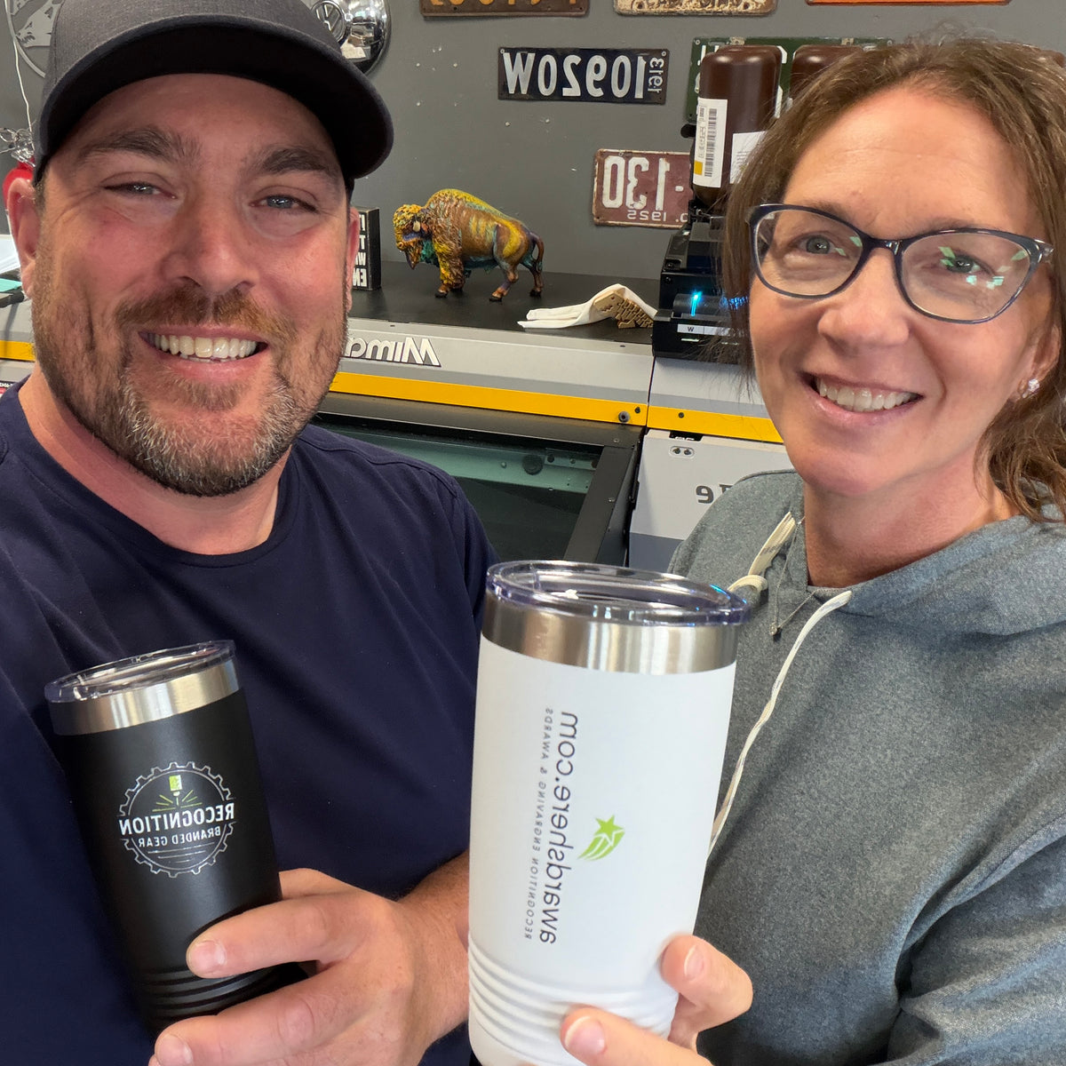 A man and a woman smiling while holding custom printed tumblers in a workshop setting. The man holds a black insulated tumbler with a silver lid, and the woman holds a white insulated tumbler with a clear lid and green logo.
