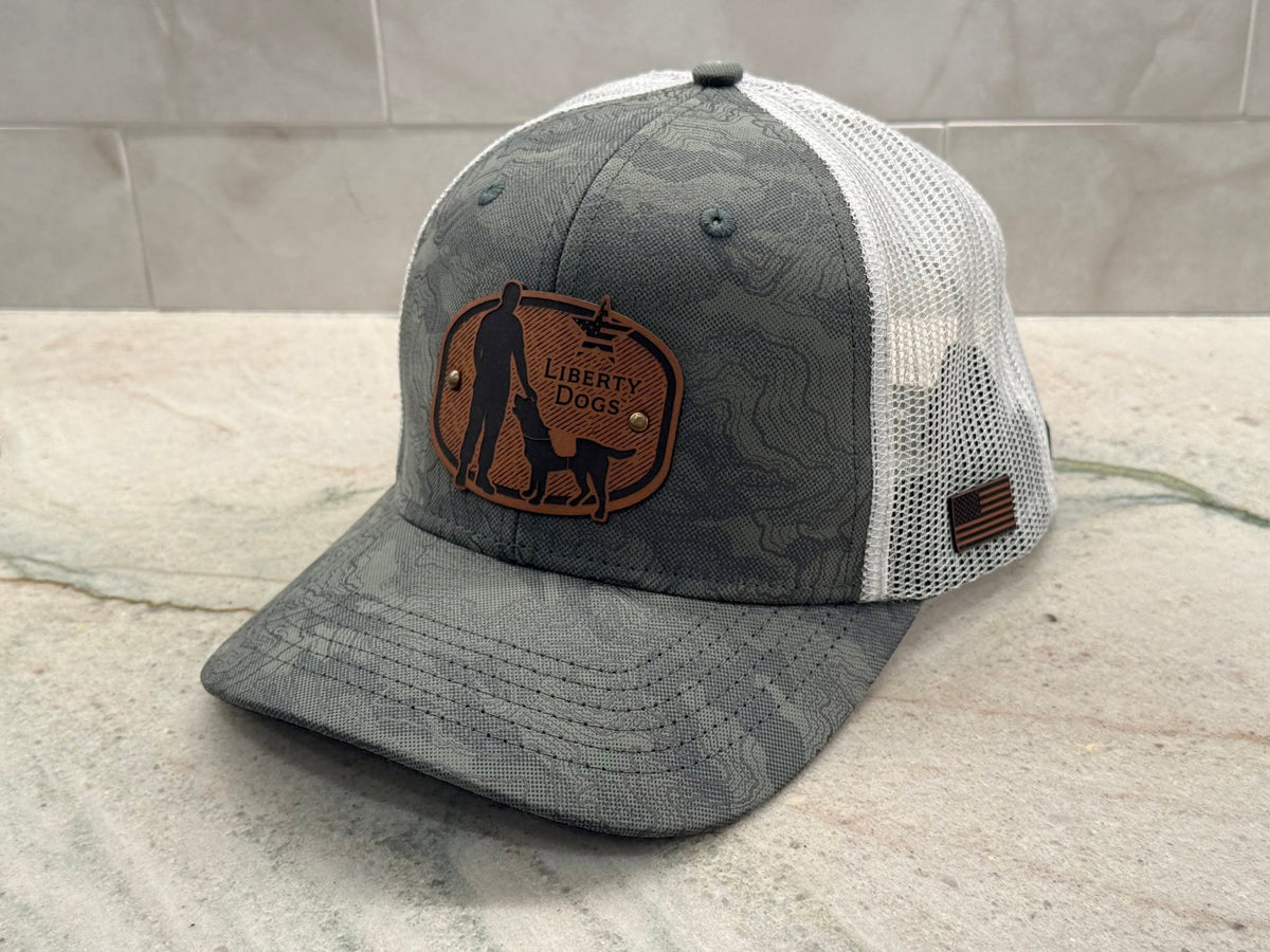 A green and white topographic patterned trucker hat with a brown leather patch on the front that reads 'Liberty Dogs'. The hat has white mesh back panels, and a small American flag patch on the side.