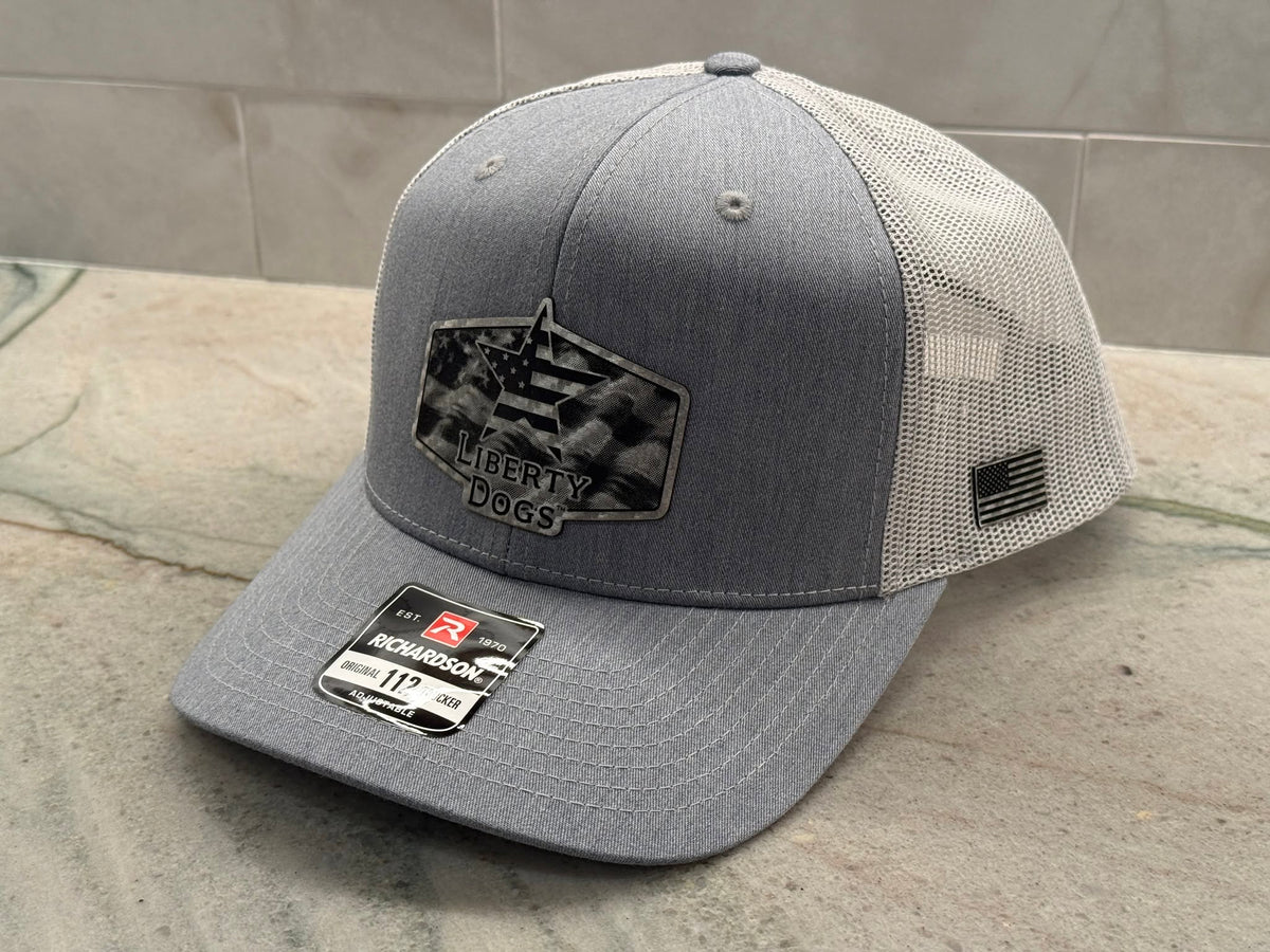 A gray and off-white trucker cap with a black and white Liberty Dogs patch on the front, resting on a stone surface.