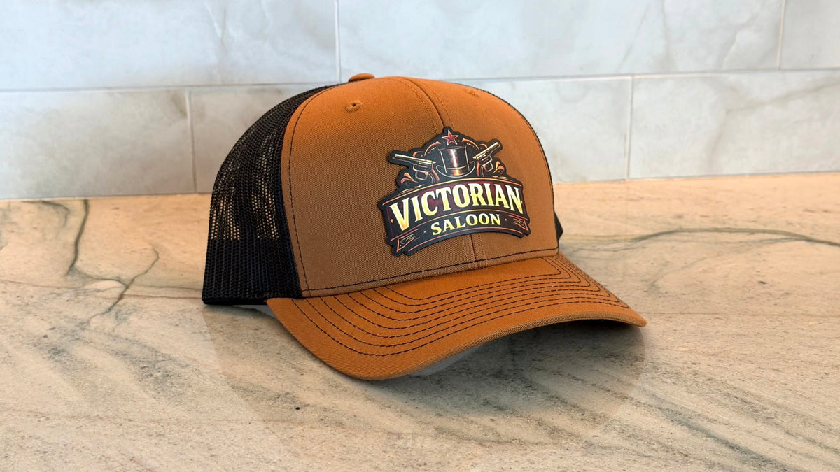 A brown and black trucker hat with a 'Victorian Saloon' color printed patch on the front, resting on a marble countertop against a tiled wall.