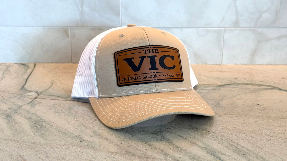 A beige and white trucker cap with a brown leather patch reading 'THE VIC - VICTORIAN SALOON • SPARKS, NV' on a marble countertop against a tiled wall.