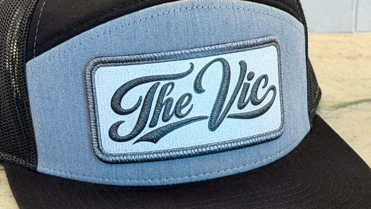 A close-up of a trucker hat featuring a light blue front panel with a rectangular UV printed embroidered look patch that reads 'The Vic' in black script. The hat has black mesh back panels and a black brim.