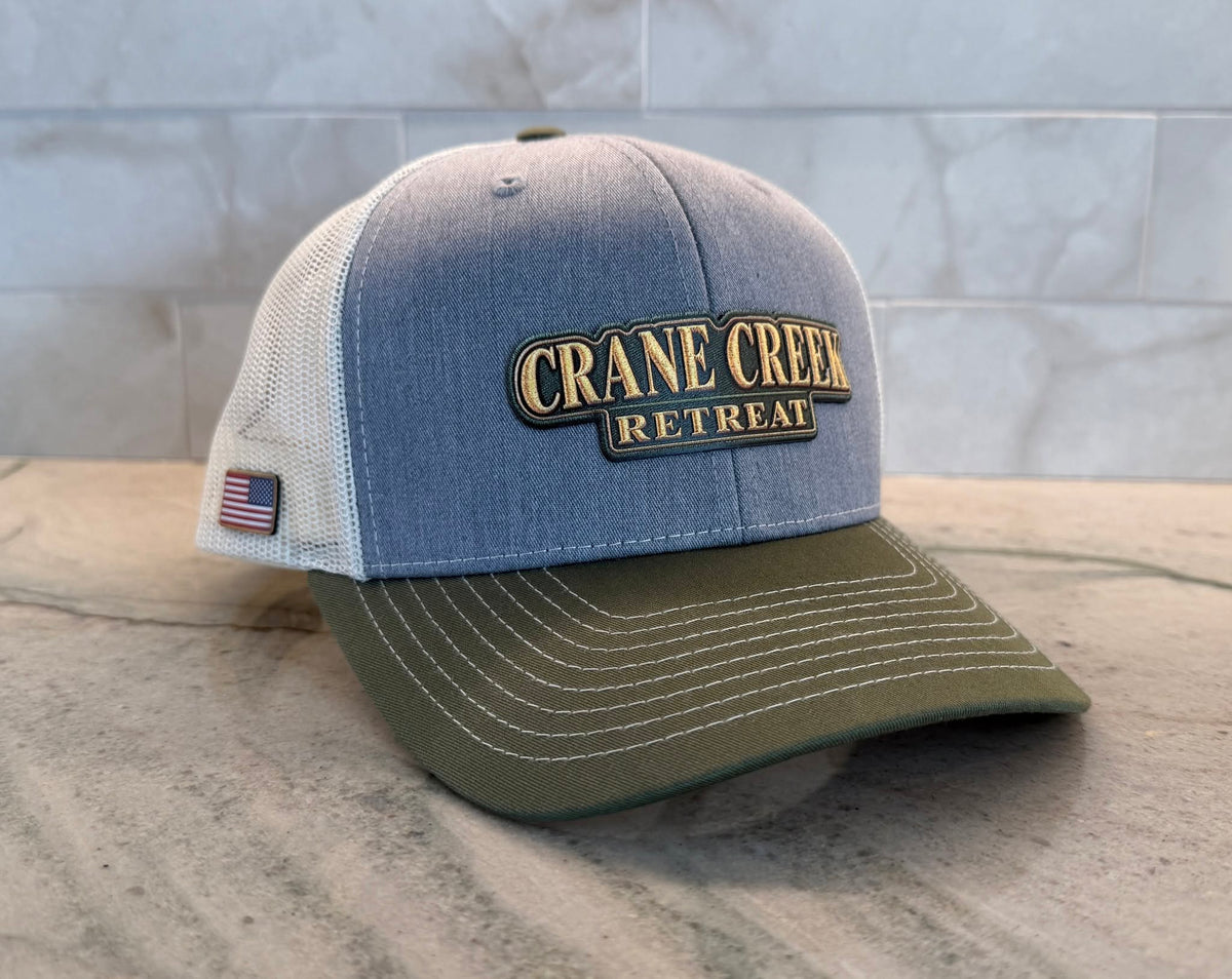 A trucker-style baseball hat with a blue-gray denim front panel, white mesh back, and olive green brim. The front features a patch reading 'CRANE CREEK RETREAT' in gold lettering, and a small American flag patch is visible on the side.