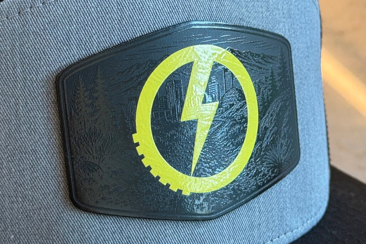 A close-up of a gray fabric cap featuring a black shield-shaped patch with a bright yellow lightning bolt and circular design with a Reno skyline in gloss clear printed over black.