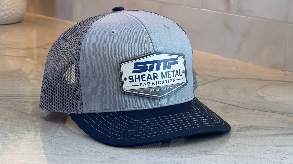A light gray and navy blue trucker hat with a mesh back, featuring a prominent white and blue 'SMF Shear Metal Fabrication' patch on the front. The hat is resting on a light gray marble surface.