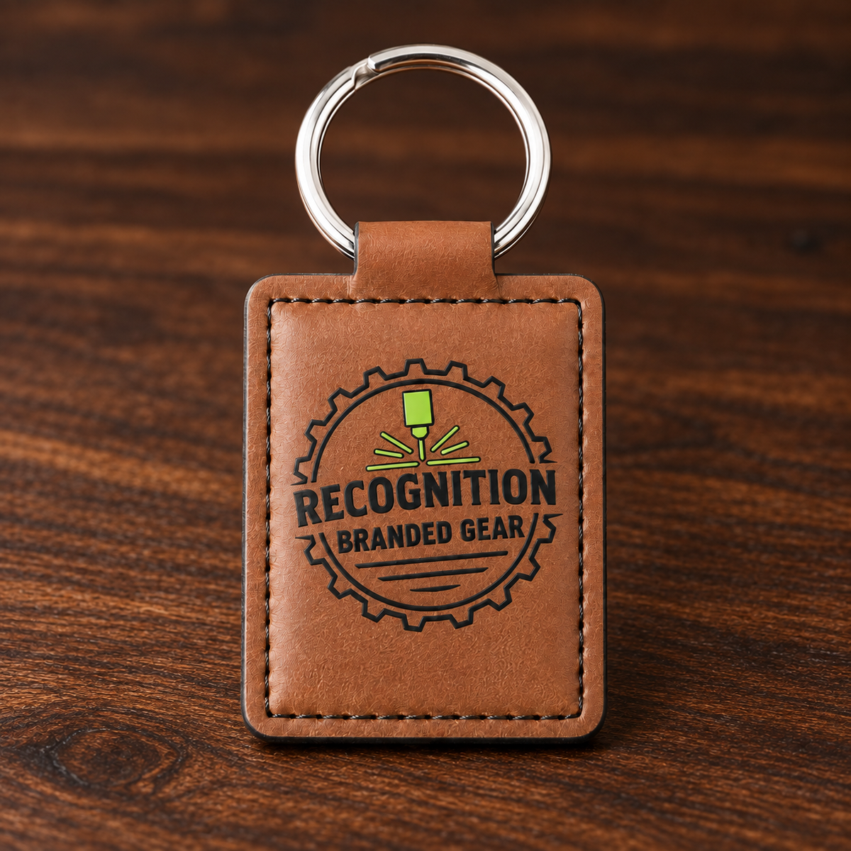 A brown leather keychain with a silver metal key ring, featuring an UV printed circular logo that reads 'RECOGNITION BRANDED GEAR' with a green and black graphic, set against a dark wood background.