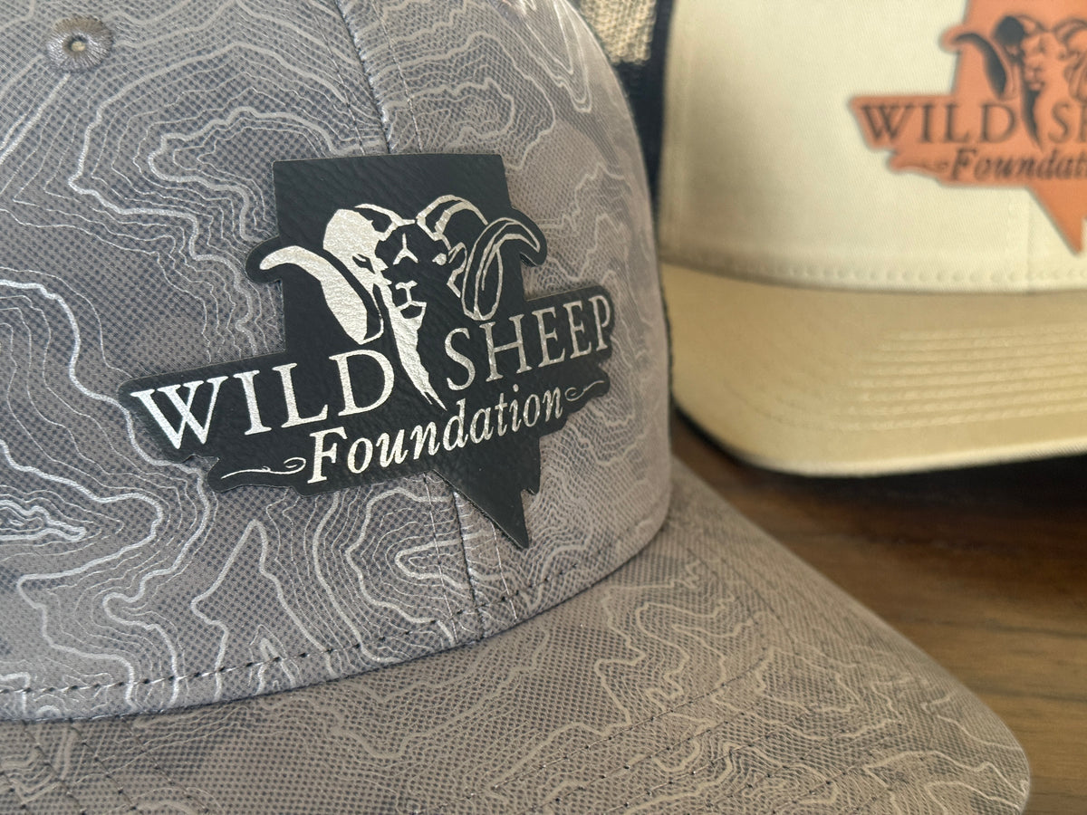 Close-up of a gray trucker hat with a topographic map pattern and a black patch logo reading 'Wild Sheep Foundation' featuring a ram illustration. A second cream-colored hat with a similar logo is partially visible in the background on a wooden surface.