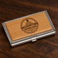 A rectangular metal business card case with a natural wood insert, engraved with a circular badge reading 'RECOGNITION BRANDED GEAR,' shown on a dark wood surface.