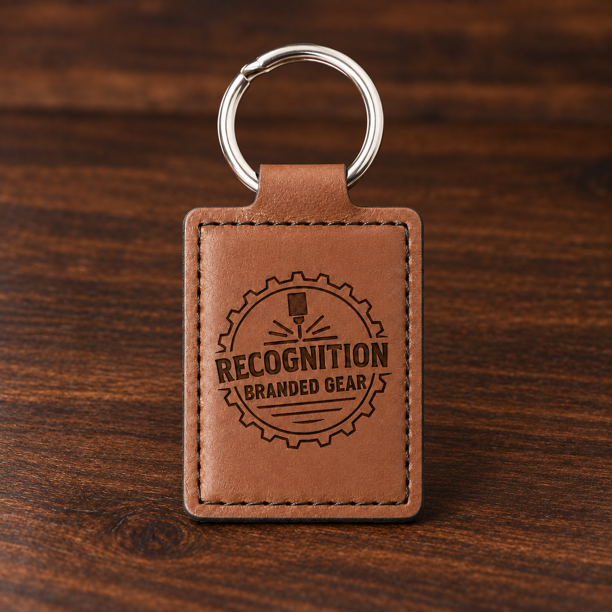 A brown leather keychain with a silver metal split ring, featuring an engraved circular badge that reads 'RECOGNITION BRANDED GEAR' on a dark wood background.