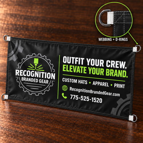A black outdoor banner with white and lime green printed text and logos, displayed on a dark wood surface. The banner has metal grommets on the corners and a circular inset diagram in the top right highlights the webbing and D-ring attachment.