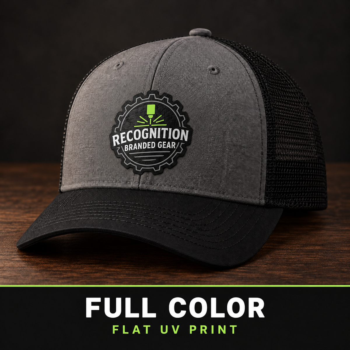 A gray and black trucker cap with a mesh back, featuring a circular embroidered patch on the front that reads 'Recognition Branded Gear' with a green and white design. The cap is displayed on a dark wooden surface against a dark background, with a black banner at the bottom reading 'FULL COLOR FLAT UV PRINT'.