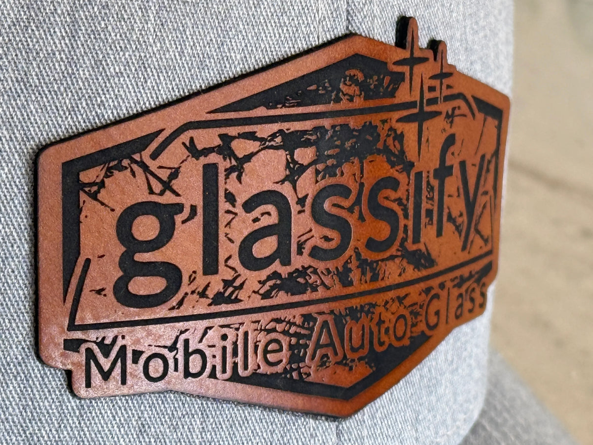 A close-up of a textured brown leather patch with black laser-cut lettering that reads 'glassify' and 'Mobile Auto Glass', attached to a gray fabric cap.