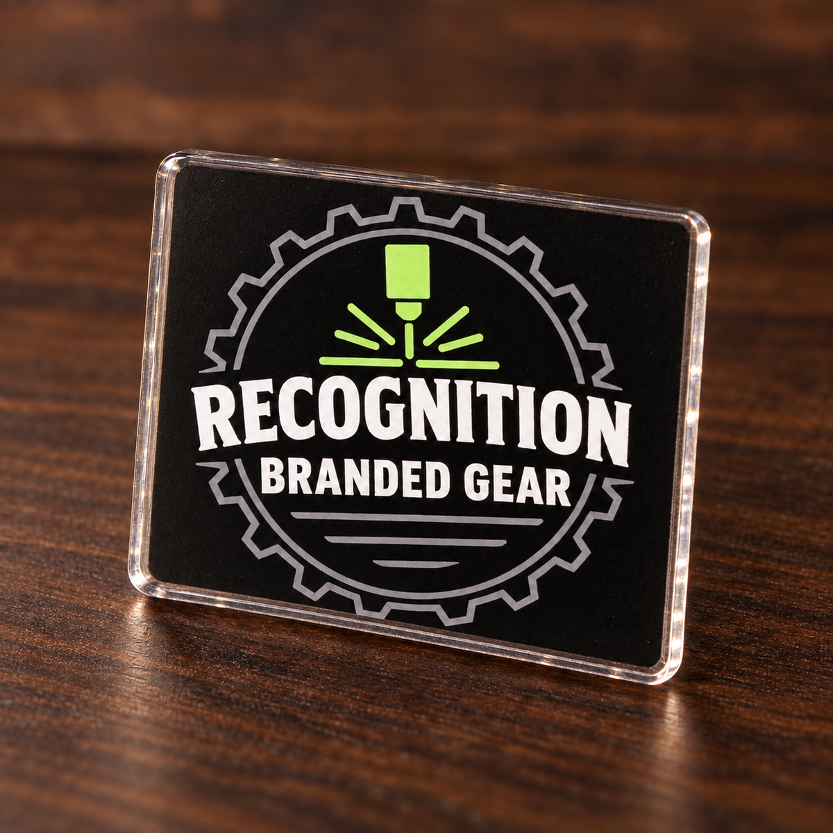 A rectangular clear acrylic refrigerator magnet standing on a dark wood surface. The design features a circular gear logo with a green laser symbol and the text 'RECOGNITION BRANDED GEAR' in white.