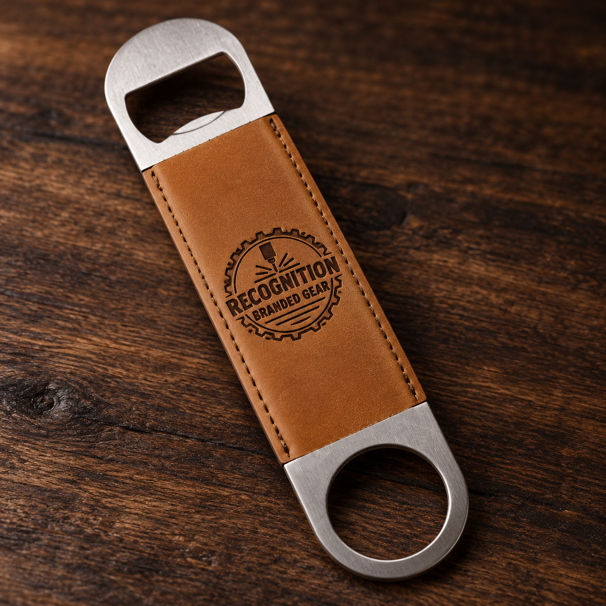 A personalized bottle opener with a brown leather handle and silver metal ends, featuring an engraved 'Recognition Branded Gear' logo, resting on a dark wood background.