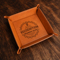 A square tan leather valet try shown on a dark brown wood surface. The inside is laser-engraved with a circular badge reading 'RECOGNITION BRANDED GEAR'.