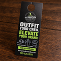 A black doorhanger promotional card for Recognition Branded Gear, photographed standing on a dark wooden surface. The card features white and bright green text reading 'OUTFIT YOUR CREW.