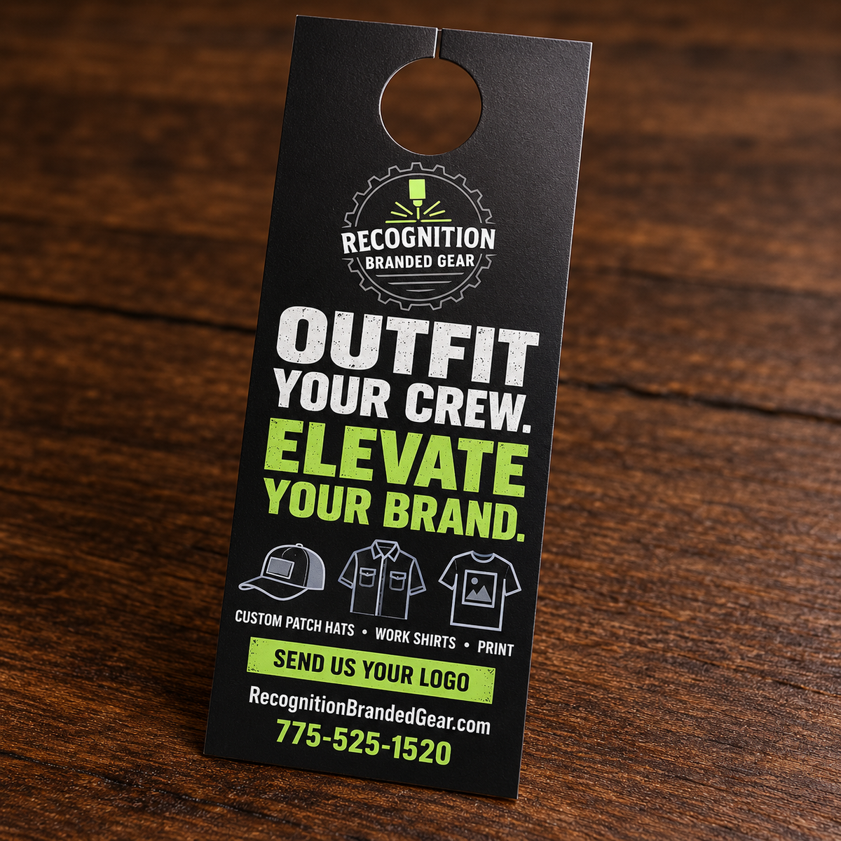 A black doorhanger promotional card for Recognition Branded Gear, photographed standing on a dark wooden surface. The card features white and bright green text reading 'OUTFIT YOUR CREW.