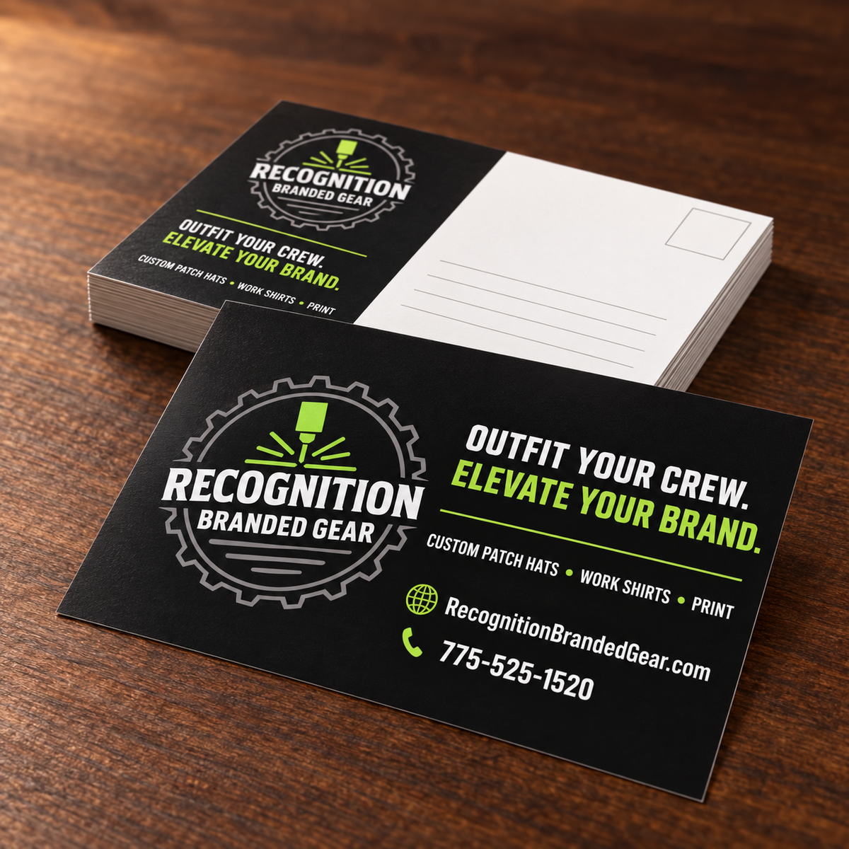A stack of custom postcards for 'Recognition Branded Gear' displayed on a dark wood tabletop. The front card is black with white and lime green text and logo, while the back card is white with a post office box design.