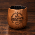 A small wooden whiskey cup with a rounded shape, featuring a laser-engraved logo that reads 'RECOGNITION BRANDED GEAR' inside a gear emblem. The tumbler is made of medium brown wood with a black interior, set on a dark wood surface.