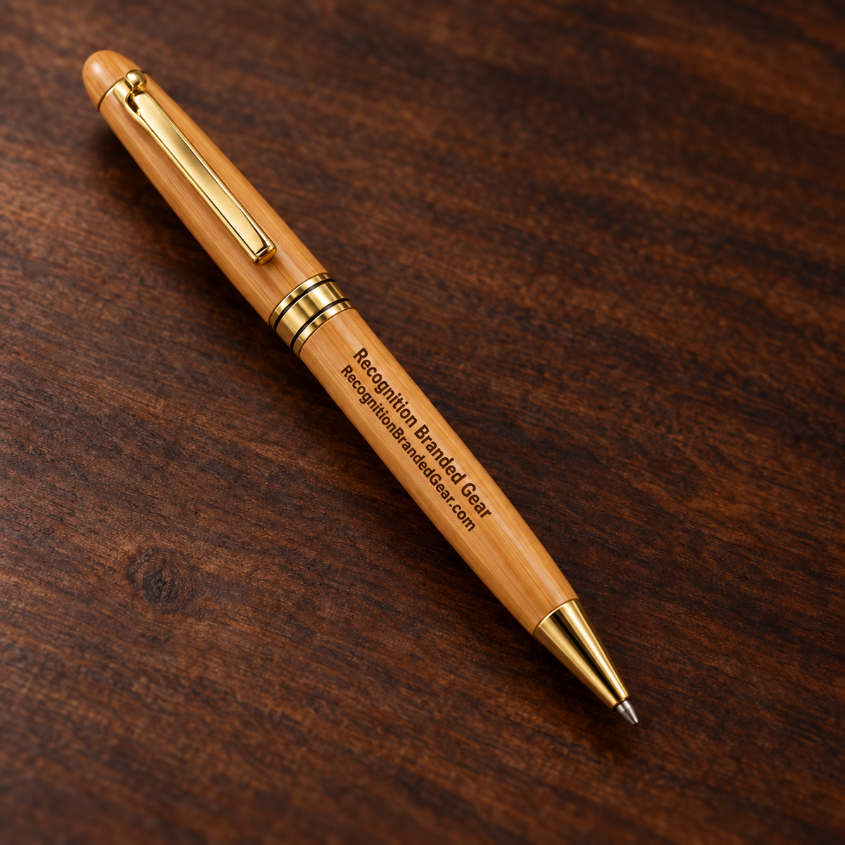 A wooden ballpoint pen with gold metal accents and custom printed text, resting diagonally on a dark brown wood grain surface.