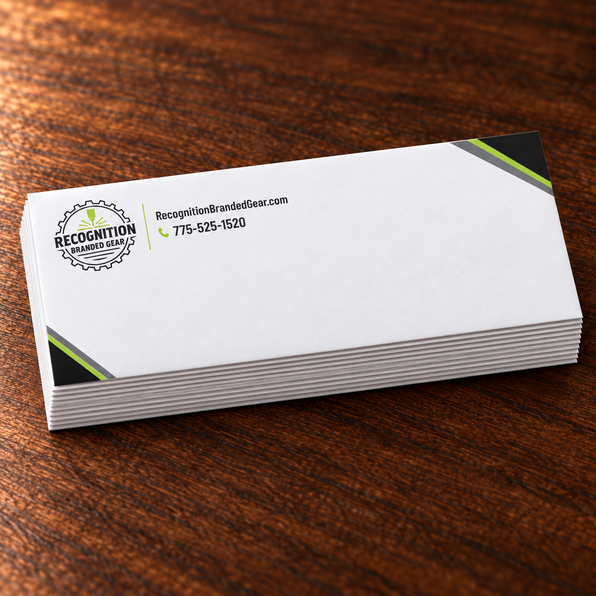 A stack of white #10 envelopes with black and green corner accents and printed logo and contact details, photographed on a dark brown wooden surface.