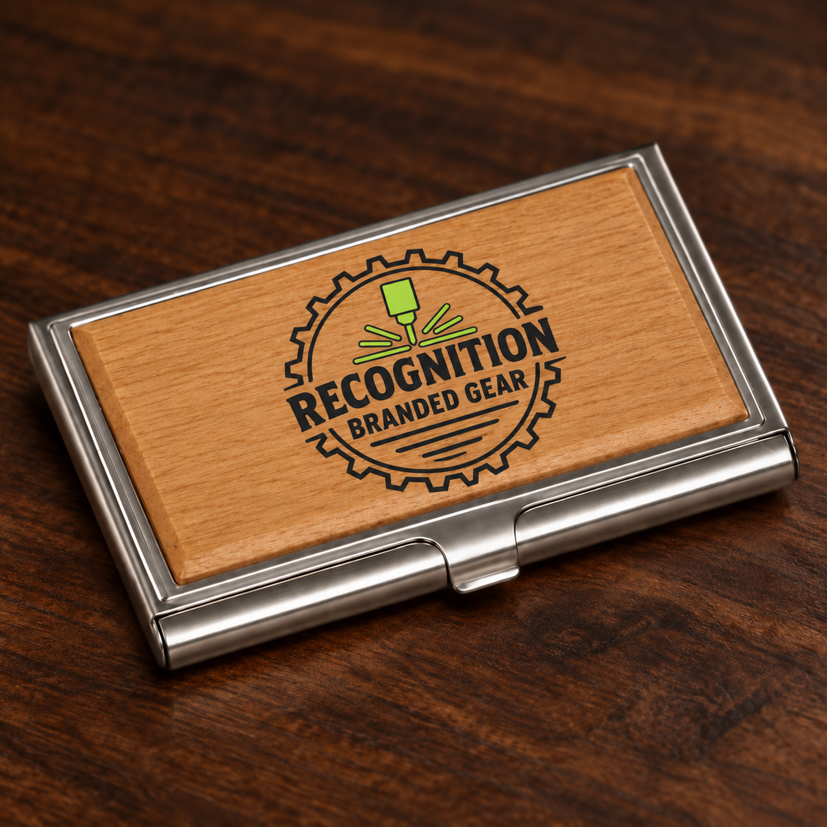 A rectangular metal business card case with a natural wood insert, photographed on a dark wood tabletop. The insert is UV printed in color with a circular gear logo and the words 'RECOGNITION BRANDED GEAR' in black, with a small green tool graphic above the text.