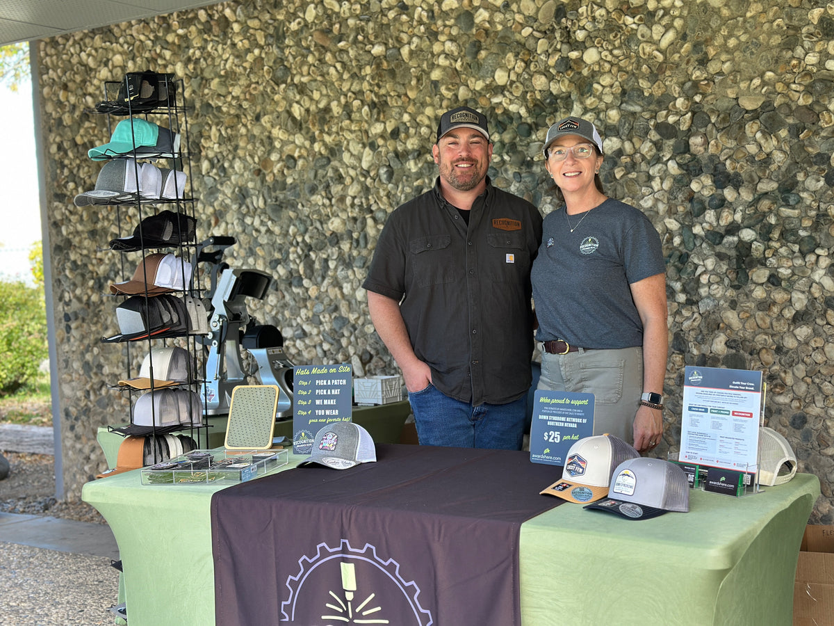 Two smiling owners stand behind a promotional table displaying baseball caps and informational signs in front of a textured stone wall.