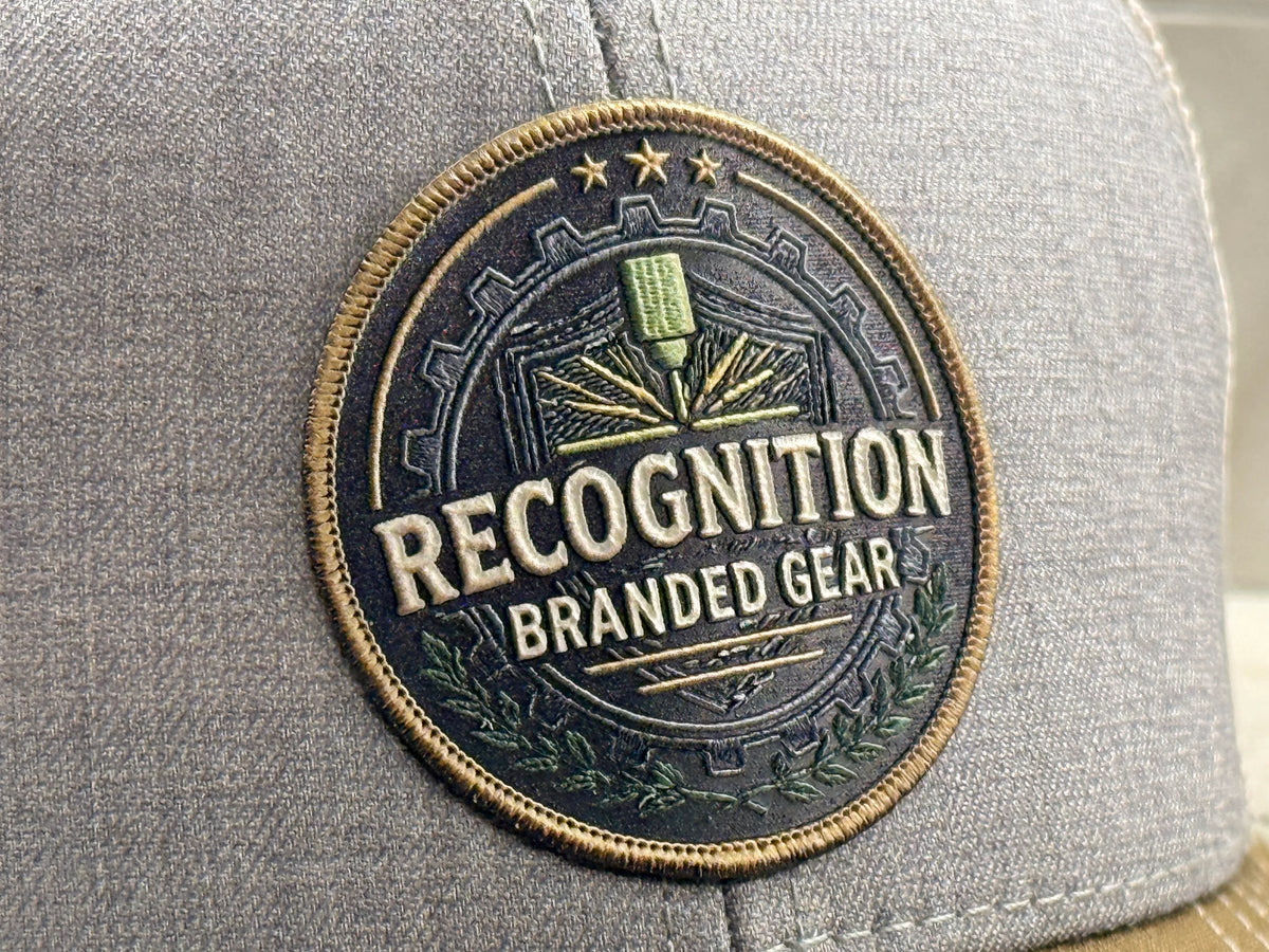 A close-up of a circular UV printed embroidered look patch on a gray fabric hat. The patch reads 'RECOGNITION BRANDED GEAR' with a laser and gear design in blue, gold, and green.