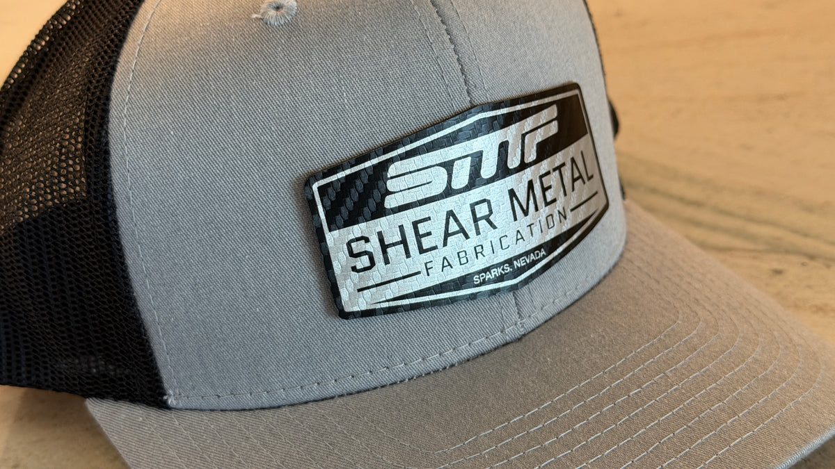 A close-up of a gray and black trucker hat with a prominent black and silver carbon fiber 'SMF Shear Metal Fabrication' patch on the front