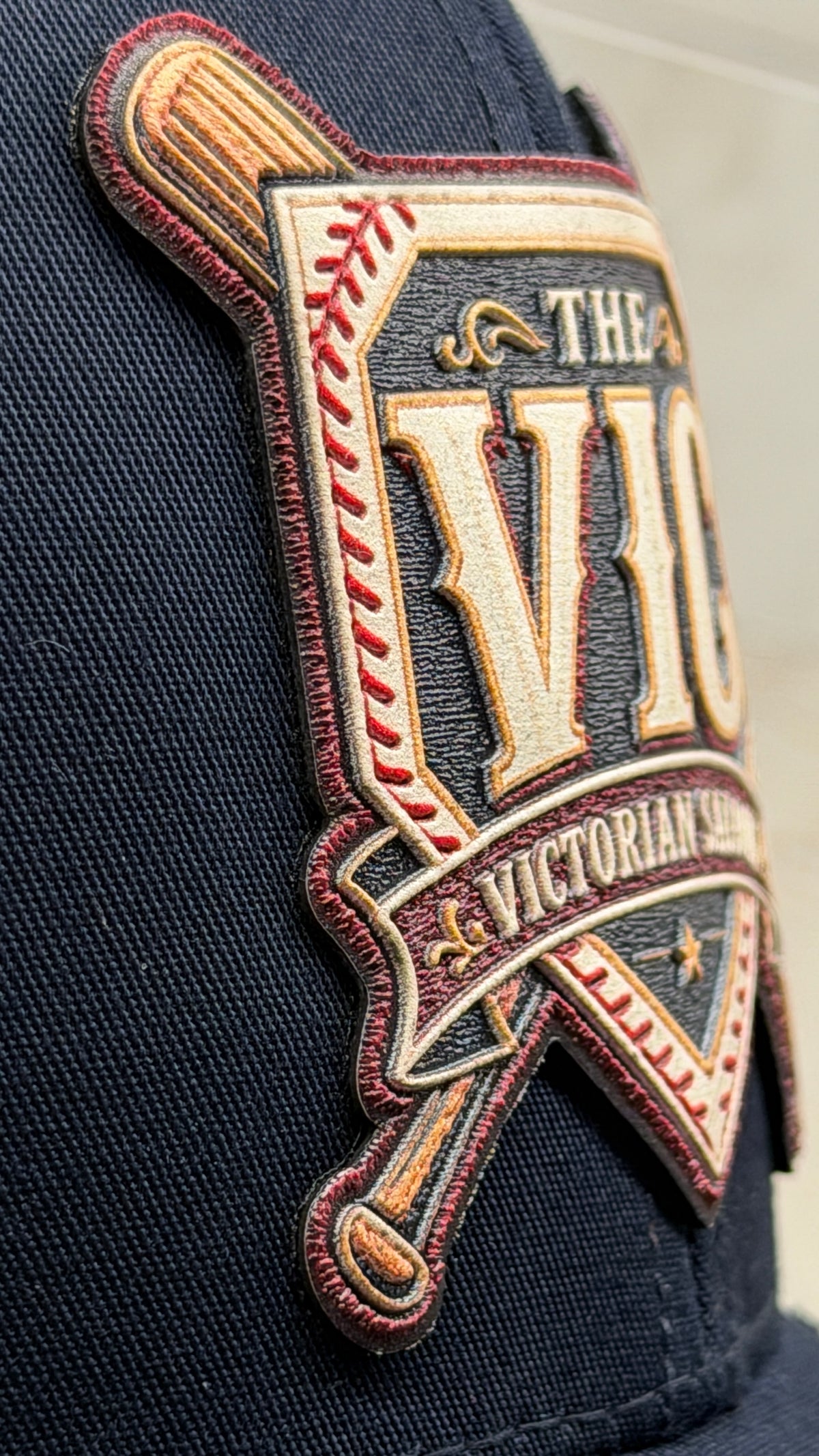 Close-up of a navy blue baseball cap featuring a large UV printed embroidery look patch with the words 'THE VIC & VICTORIAN SALOON' and crossed bats.