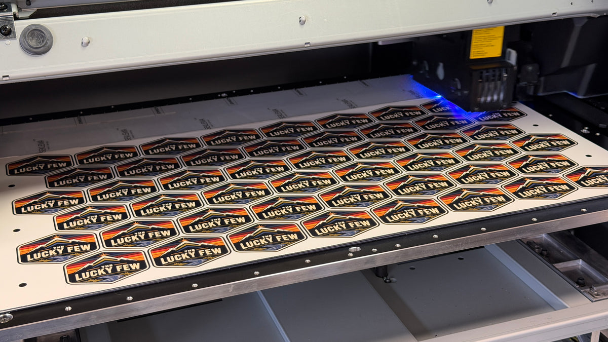 A close-up of a large industrial UV printer  with a bed of small blue, orange, red, grean and white 'LUCKY FEW' shield-shaped patches being processed under a blue light.