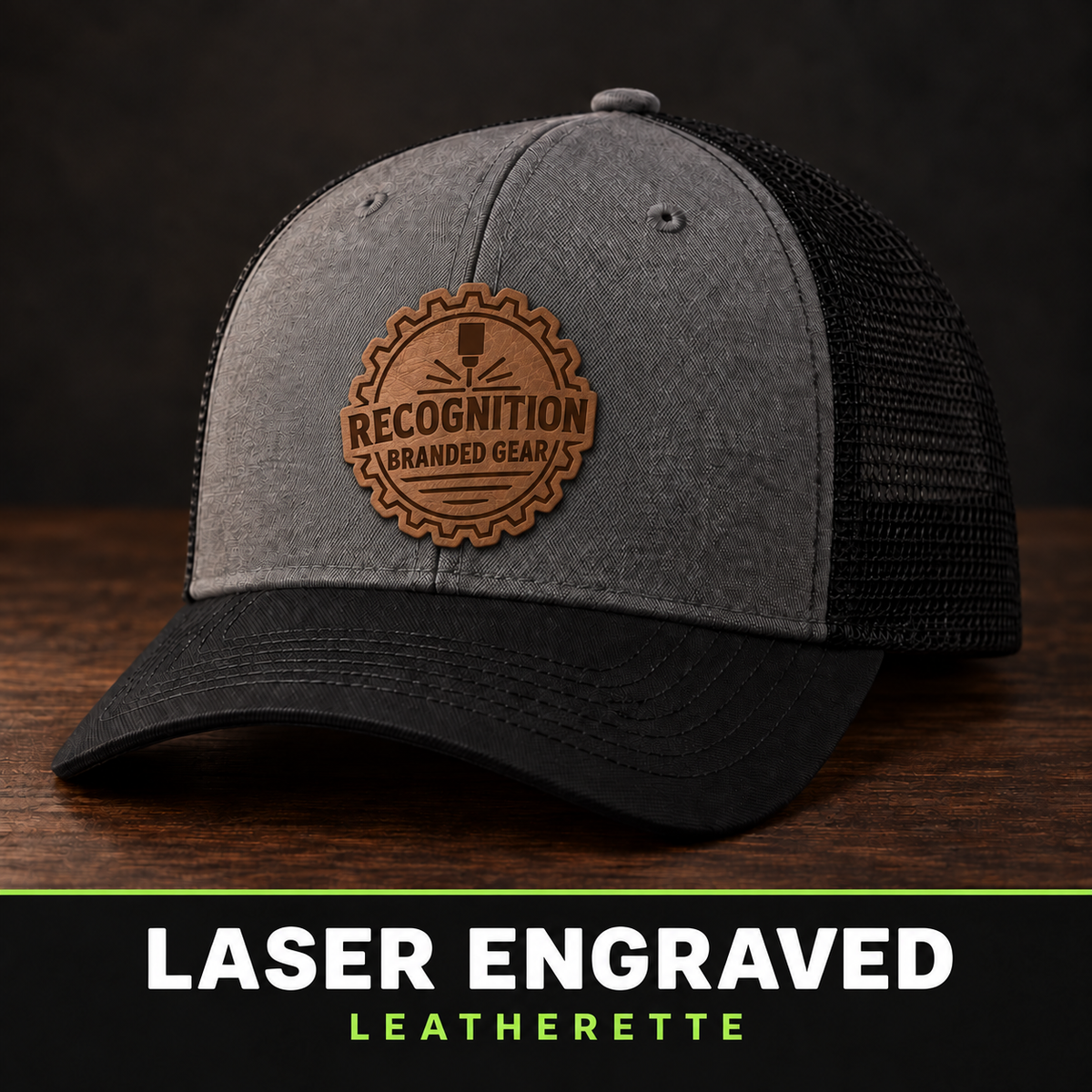 A gray and black trucker cap with a brown leatherette patch engraved with 'Recognition Branded Gear' on the front, displayed on a dark wooden surface against a dark background with a text banner at the bottom reading 'LASER ENGRAVED LEATHERETTE'.
