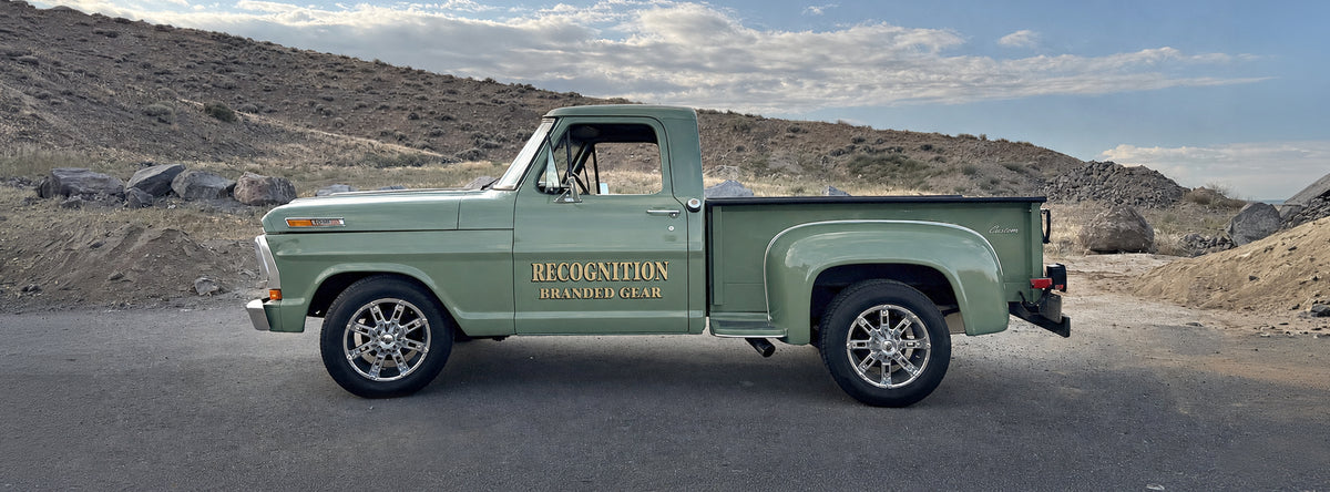 A vintage green Ford pickup truck parked outdoors on a paved road with rocky hills and a blue sky in the background. The side of the truck has gold lettering that reads 'RECOGNITION BRANDED GEAR'.
