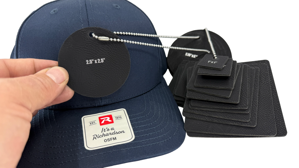 A navy blue baseball cap with a white and red Richardson 0SFM patch on the front, shown alongside black leather hang tags and sizing cards. A hand is holding one of the black leather hang tags on a silver ball chain against a plain white background.