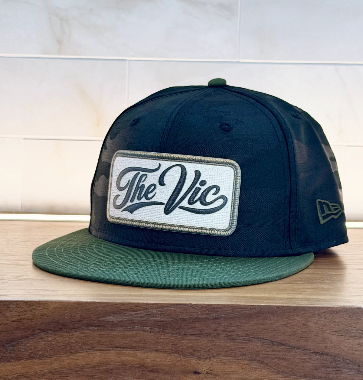 A dark navy blue snapback cap with a green visor and a white rectangular patch on the front that reads 'The Vic' in black script. A small New Era flag logo is visible on the side.
