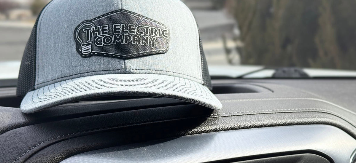 A gray heather trucker cap with a black mesh back and a black embroidered patch reading 'THE ELECTRIC COMPANY' sits on the dashboard of a vehicle.