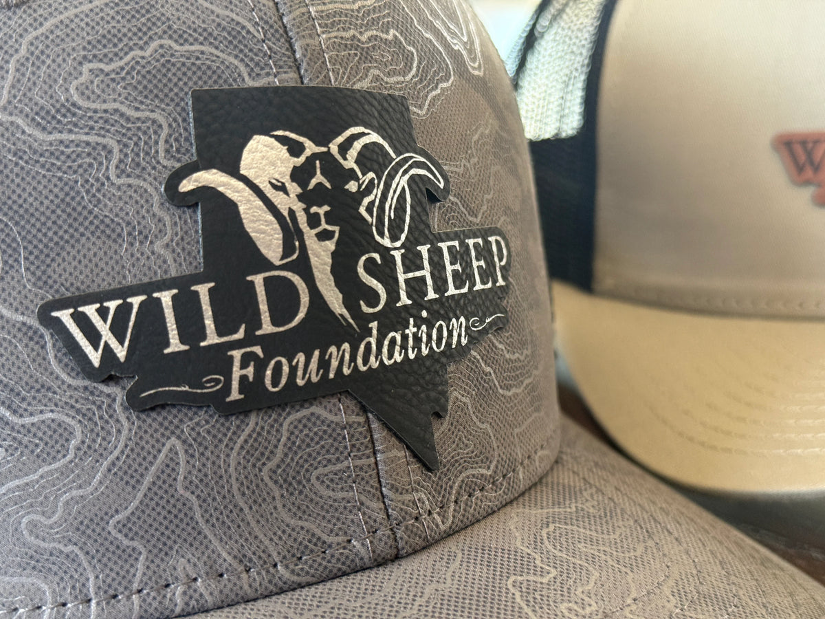 Close-up of a gray patterned trucker cap with a black patch reading 'Wild Sheep Foundation' and a ram graphic, with another cap partially visible in the background.